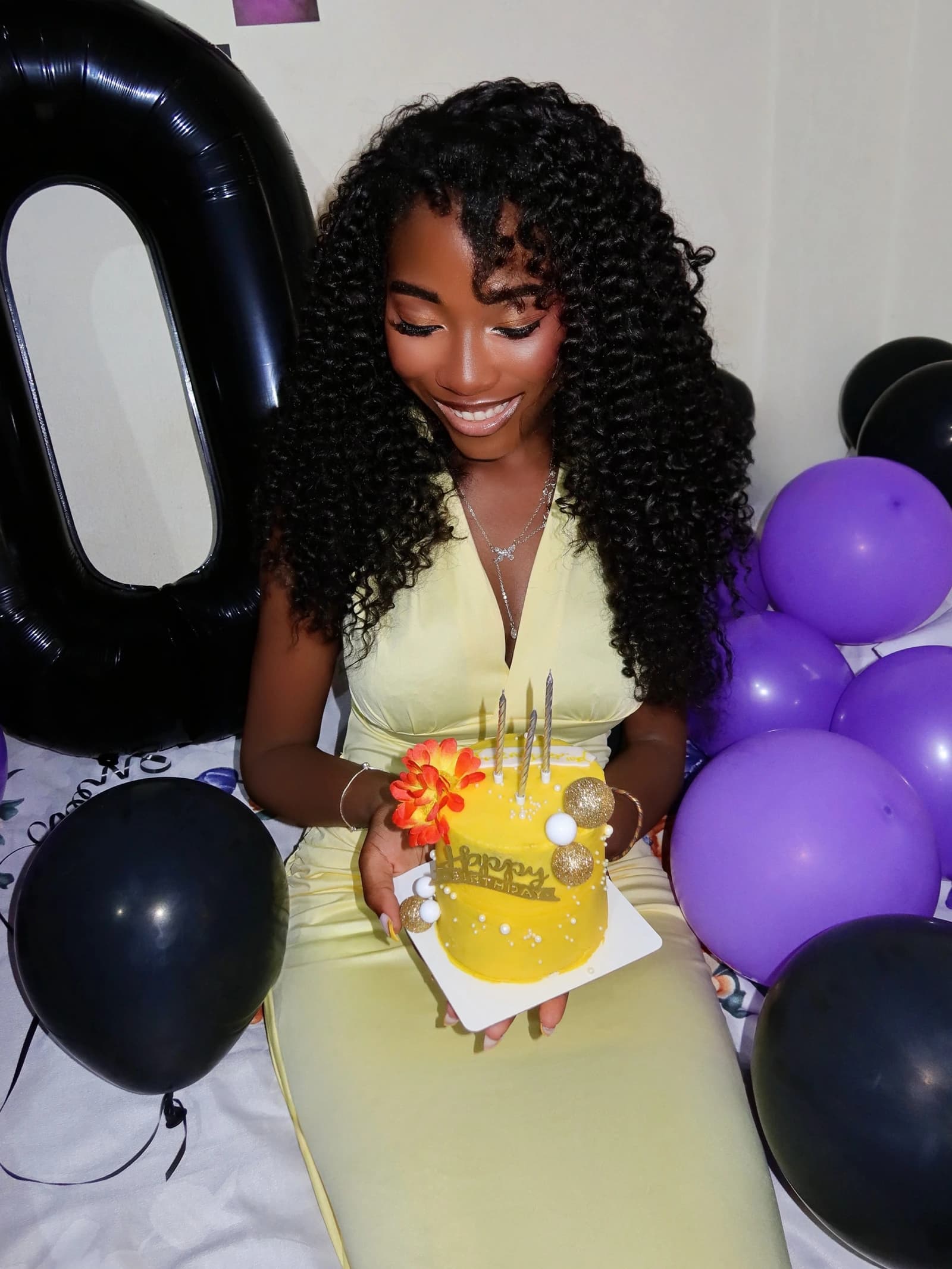 Smiling portrait looking down at a yellow birthday cake, surrounded by purple and black balloons.