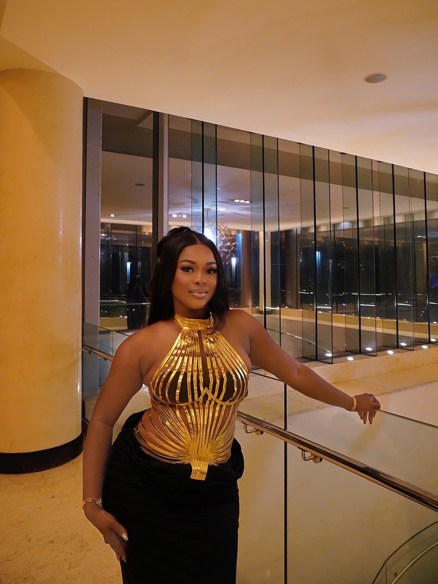 Woman in a sculptural gold halter and black skirt beside a mirrored staircase.