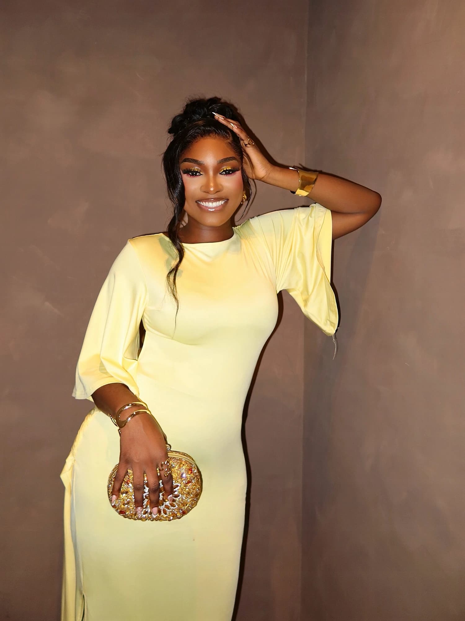 Front portrait in a pale yellow dress with a gold clutch and one hand in the hair.